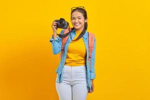 portrait d'une jeune femme asiatique souriante en vêtements en denim avec sac à dos et montrant un appareil photo professionnel isolé sur fond jaune
