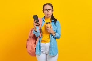 portrait d'une jeune étudiante asiatique agacée en vêtements denim avec sac à dos tenant une tasse de café et smartphone isolé sur fond jaune photo