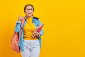 portrait d'une jeune étudiante asiatique souriante en vêtements en denim avec sac à dos tenant un cahier et faisant un geste téléphonique comme dit rappelez-moi isolé sur fond jaune photo