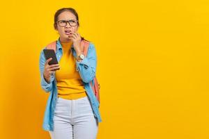 portrait d'une jeune étudiante asiatique sérieuse en vêtements décontractés avec sac à dos à l'aide d'un téléphone portable et se mordant les ongles tout en regardant de côté isolé sur fond jaune photo