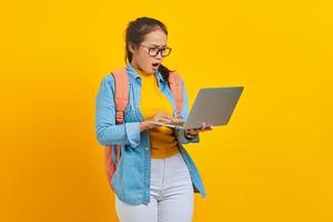 portrait d'une jeune étudiante asiatique surprise en vêtements décontractés avec sac à dos regardant les e-mails entrants sur un ordinateur portable isolé sur fond jaune. éducation au concept de collège universitaire photo