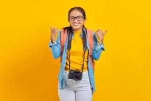 joyeuse jeune femme asiatique voyageuse avec sac à dos et appareil photo dans des vêtements en denim tout en montrant le geste du pouce vers le haut isolé sur fond jaune. passager voyageant le week-end. concept de voyage en avion