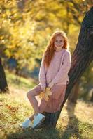 portraits d'une charmante fille rousse avec un joli visage. fille posant dans le parc d'automne dans un pull et une jupe de couleur corail. dans les mains d'une fille une feuille jaune photo