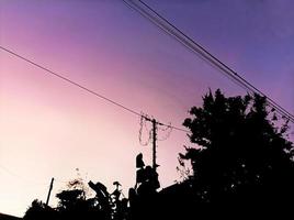 photo en silhouette de maisons, de poteaux électriques et de plantes à l'aube dans le ciel bleu, violet et rose du village.