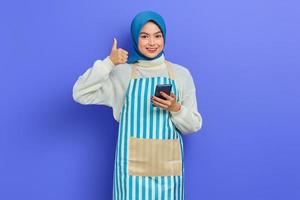 portrait d'une jeune femme musulmane asiatique souriante portant le hijab et un tablier à l'aide d'un smartphone, gesticulant les pouces vers le haut isolés sur fond violet. concept de mode de vie musulman femme au foyer photo