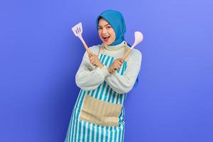 joyeuse belle femme asiatique en pull blanc recouvert de tablier et hijab main croisée tenir la spatule et la cuillère tout en faisant le ménage isolé sur fond violet. notion de ménage photo