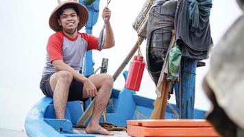 heureux jeune pêcheur sur la plage tenant son poisson et montre devant son bateau photo