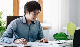 portrait d'un jeune homme asiatique pigiste à la main travaille son travail sur une tablette informatique dans un bureau moderne. faire un rapport d'analyse comptable sur les données d'investissement immobilier, le concept de systèmes financiers et fiscaux. photo