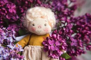 petit ange aux cheveux d'or dans les fleurs de lilas bleu, rose, violet, violet. jouet fait à la main dans des couleurs lilas violettes. carte de voeux.