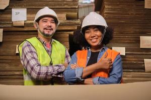 travailleuse afro-américaine en uniforme de sécurité et casque avec un collègue asiatique masculin regardant la caméra et le sourire, inspecteur de l'entrepôt de l'usine de l'industrie, piles de fabrication de carton empilable. photo