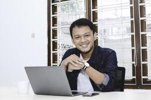 jeune homme asiatique se sentant heureux et souriant quand il travaille sur un ordinateur portable sur la table. homme indonésien portant une chemise bleue. photo