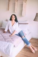 portrait d'une belle femme aux cheveux noirs en chemise blanche et jeans assis triste sur son lit. émotions, crise, maison, concept d'intérieur