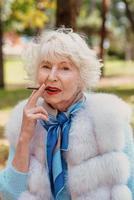 femme senior élégante et élégante souriante à la mode avec des cheveux gris en manteau de fourrure cigarette en plein air. mode de vie malsain, âge, vibrations positives, vieillesse, dépendance, concept de mauvaise habitude. photo