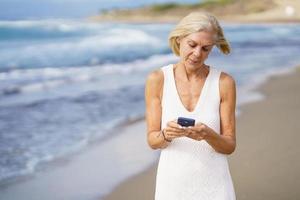 femme âgée marchant sur la plage à l'aide d'un smartphone. photo