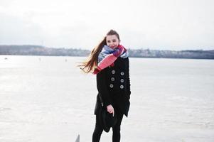 jeune fille décontractée au manteau noir, écharpe et chapeau contre la rivière gelée par temps d'hiver ensoleillé. photo
