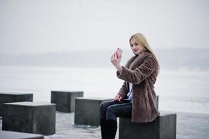 Élégante jeune fille blonde riche en manteau de fourrure regardant sur un smartphone rose sur des cubes de pierre de fond à la main contre un lac gelé le jour de l'hiver. photo