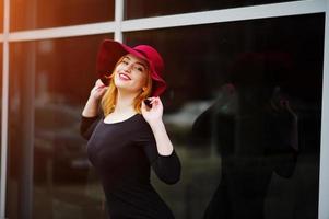 portrait d'une fille aux cheveux rouges fashion sur un chapeau rouge et une robe noire avec un maquillage lumineux posé contre une grande fenêtre. filtres instagram de style photo tonique.