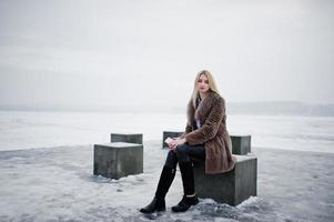 Élégante jeune fille blonde riche en manteau de fourrure avec un smartphone rose sur des cubes de pierre de fond à la main contre un lac gelé le jour de l'hiver. photo