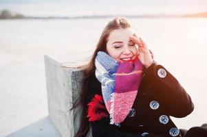 jeune fille décontractée au manteau noir, écharpe et chapeau contre la rivière gelée par temps d'hiver ensoleillé. photo