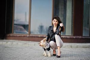 brune gitane avec chien yorkshire terrier posé contre de grandes fenêtres maison. le modèle porte une veste en cuir et un t-shirt avec ornement, un pantalon et des chaussures à talons hauts. photo