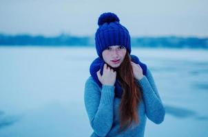 portrait d'une jeune fille aux cheveux roux avec des taches de rousseur portant un bonnet et une écharpe en laine tricotée bleue dans la glace de fond de la journée d'hiver. photo