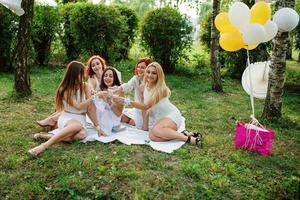 cinq filles avec du champagne à portée de main portées sur une robe blanche lors d'une fête de poule assise en plein air au parc et s'amusant. photo