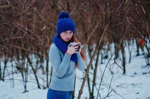 portrait de jeune fille aux cheveux roux avec des taches de rousseur portant un bonnet et une écharpe en laine tricotée bleue avec une tasse de thé en journée d'hiver. photo