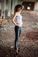 portrait d'une jeune fille brune mignonne portant un pantalon en cuir noir et un chemisier blanc posé sur un lieu abandonné. photo