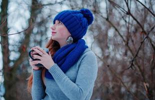 portrait de jeune fille aux cheveux roux avec des taches de rousseur portant un bonnet et une écharpe en laine tricotée bleue avec une tasse de thé en journée d'hiver. photo