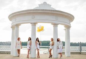 cinq filles avec des ballons à portée de main portées sur des robes blanches lors d'une fête de poule contre des colonnes blanches d'arc. photo