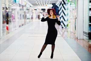 mode fille aux cheveux rouges portant une robe noire et un chapeau rouge posés au centre commercial du commerce. filtres instagram de style photo tonique.