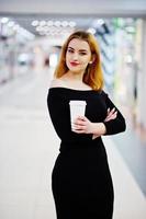mode fille aux cheveux rouges porter sur une robe noire avec un maquillage lumineux tenant une tasse de café au centre commercial commercial. filtres instagram de style photo tonique.