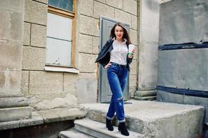 portrait d'une jeune fille élégante portant une veste en cuir et un jean déchiré avec une tasse de café. style de modèle de mode de rue. photo