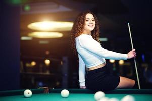 jeune fille frisée posée près de la table de billard. modèle sexy à mini mini jupe noire jouer au billard russe. jouer au jeu et au concept amusant. photo