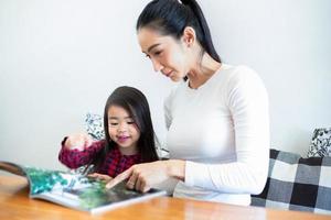 une mère asiatique apprend à sa fille à lire un livre pendant la pause semestrielle sur la table de salon et à avoir du lait froid sur la table à la maison. concepts éducatifs et activités de la famille photo