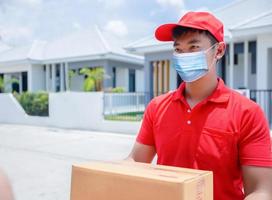 des livreurs asiatiques portant un uniforme rouge avec une casquette rouge et un masque facial manipulant des boîtes en carton à donner à la cliente devant la maison. achats en ligne et livraison express photo