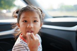petite jeune fille asiatique restant dans la voiture et mangeant des chips de pommes de terre frites, une jolie fille aime manger une collation tout en voyageant en voiture. photo