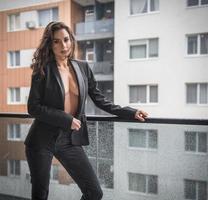 magnifique femme brune glamour avec une veste noire posant sur un balcon moderne avec une vue imprenable sur la ville. portrait d'une femme élégante à la mode avec de longues jambes, un jean noir portant un balcon photo