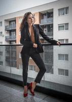 magnifique femme brune glamour avec une veste noire posant sur un balcon moderne avec une vue imprenable sur la ville. portrait d'une femme élégante à la mode avec de longues jambes, un jean noir portant un balcon photo