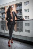 magnifique femme brune glamour avec une veste noire posant sur un balcon moderne avec une vue imprenable sur la ville. portrait d'une femme élégante à la mode avec de longues jambes, un jean noir portant un balcon