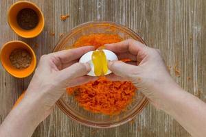 les mains de la femme mettent l'oeuf cru de l'oeuf cassé dans la pâte de carottes râpées pour la tarte aux carottes sur le fond en bois photo