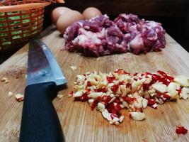 nom des ingrédients de cuisine boeuf au basilic sauté avec un œuf au plat. et les piments frais hachés avec de l'ail placent sur une planche à découper en bois avec un couteau. feuilles de basilic sacré dans un panier orange photo