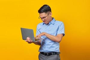 portrait d'un jeune homme asiatique bouleversé asiatique dans des verres à l'aide d'un ordinateur portable isolé sur fond jaune. concept d'homme d'affaires et d'entrepreneur photo