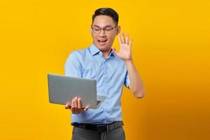 portrait d'un jeune homme asiatique joyeux asiatique dans des verres à l'aide d'un ordinateur portable, faisant un appel vidéo, faisant signe de salut à des amis isolés sur fond jaune. concept d'homme d'affaires et d'entrepreneur photo
