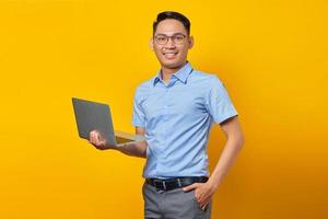 portrait d'un bel homme asiatique souriant dans des verres tenant un ordinateur portable et regardant la caméra isolée sur fond jaune. concept d'homme d'affaires et d'entrepreneur photo