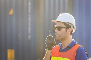 un contremaître portant un casque et un gilet de sécurité parle sur une radio bidirectionnelle, un ouvrier d'usine contrôle le chargement d'une boîte de conteneurs à partir d'une cargaison photo