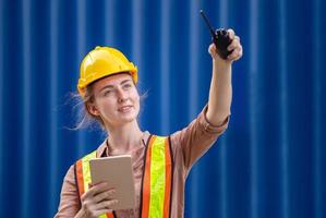 femme ingénieure portant un casque et un gilet de sécurité tenant une tablette et une radio bidirectionnelle pointant vers le ciel, ouvrière d'usine à la cargaison de conteneurs photo