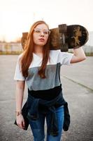 jeune adolescente urbaine avec planche à roulettes, porter des lunettes, une casquette et un jean déchiré sur le terrain de sport de la cour au coucher du soleil. photo