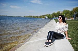 fille brune sur pantalon en cuir pour femmes et chemisier blanc, lunettes de soleil, assis sur le quai du lac. photo