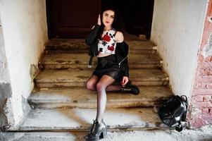 jeune fille goth sur jupe en cuir noir et veste avec sac à dos posé sur les escaliers de la vieille maison. photo